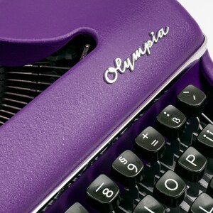 May include: A purple vintage typewriter with the word "Olympia" in silver lettering on the top. The typewriter has a black keyboard with keys labelled with letters, numbers, and symbols.