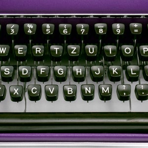 May include: A purple typewriter with a black keyboard and silver accents. The keyboard has the standard letter keys, numbers, and symbols. The typewriter has a black carriage return lever and a black space bar.