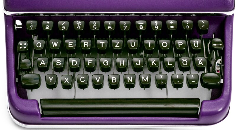 May include: A purple typewriter with a black keyboard and silver accents. The keyboard has the standard letter keys, numbers, and symbols. The typewriter has a black carriage return lever and a black space bar.