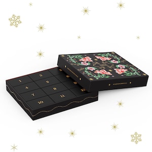 May include: Black advent calendar with 12 numbered compartments. The lid features a floral design with pink and green accents. The text "12 Days of Christmas" is visible on the lid. Gold snowflakes are scattered around the calendar.