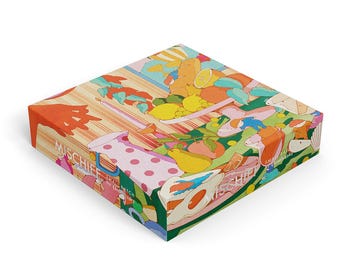 Op de afbeelding: Een vierkante doos met een kleurrijke, grillige illustratie van een picknickscène. De illustratie toont fruit, eten en een tafelkleed met stippen. Het woord "MISCHIEF" staat op de zijkant van de doos.