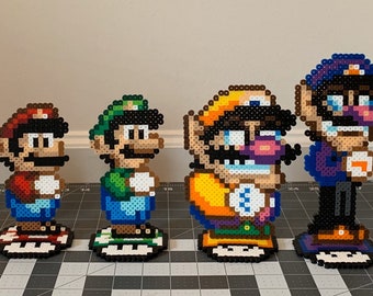 May include: A collection of eight pixel art figures of characters from the Super Mario franchise. The figures are made of colorful beads and are standing on small platforms. The characters include Mario, Luigi, Toad, Peach, Bowser, Wario, Waluigi, and Daisy.