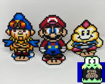 May include: Three pixel art figures of video game characters: a character in blue and gold armor, Mario in his classic red and blue outfit, and a yellow character. Each figure is on a white base. A logo in the bottom right corner reads "Pixel Dino Crafts".