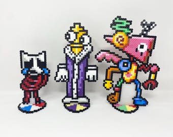 May include: Three pixel art figures made of colorful beads. The figures are standing on circular bases. The figure on the left is a white and red cat with a sad expression. The figure in the middle is a purple and yellow figure wearing a crown. The figure on the right is a pink and yellow figure with a green leafy crown and a red and yellow body.
