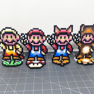 Könnte beinhalten: Vier Pixel-Art-Figuren aus bunten Perlen. Die Figuren sind Mario, Luigi, Toad und Donkey Kong. Die Figuren stehen auf einer schwarzen und grauen Oberfläche.