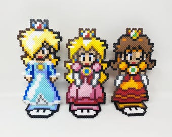 Może przedstawiać: Trzy figurki pixel art postaci z gier wideo. Figurki wykonane są z kolorowych plastikowych koralików. Postacie mają na sobie suknie i korony. Lewa postać ma na sobie niebieską suknię, środkowa postać różową suknię, a prawa postać czerwoną suknię.