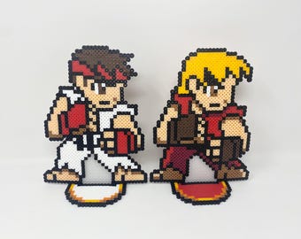May include: Two pixel art figures of video game characters, one with red and white clothing and a red headband, the other with red and brown clothing and blonde hair. Both figures are standing on a base with a pixelated design.