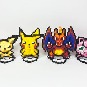 Könnte beinhalten: Vier farbenfrohe Pixel-Art-Figuren beliebter Charaktere. Die Figuren umfassen ein gelbes Pikachu, ein rotes und orangefarbenes Glurak und ein rosa Pummeluff. Jede Figur besteht aus kleinen, farbigen Perlen.