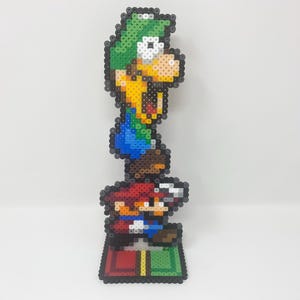 May include: A pixel art figure of Luigi standing on top of Mario, who is standing on a green and red platform. Both figures are made of colorful plastic beads.