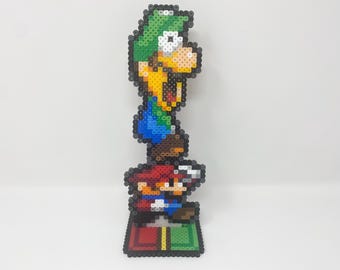May include: A pixel art figure of Luigi standing on top of Mario, who is standing on a green and red platform. Both figures are made of colorful plastic beads.