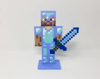 Puede incluir: Un personaje de Minecraft azul y blanco hecho de cuentas Perler, con armadura de diamante y sosteniendo una espada azul. El personaje está de pie sobre una base azul.