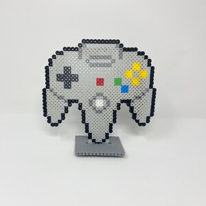 Puede incluir: Un modelo de arte de píxeles gris y negro de un mando de Nintendo 64 en un soporte gris. El mando tiene un cuerpo gris con botones negros y botones de cara amarillos, verdes, azules y rojos.