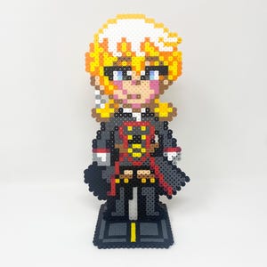 Peut inclure: Une figurine en pixel art réalisée avec des perles à repasser, représentant un personnage aux cheveux jaunes et blancs en pointe, aux yeux bleus et aux joues roses. Il porte un manteau gris foncé avec des détails rouges et jaunes sur un socle.