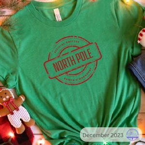 May include: A green t-shirt with a red stamp design that reads "NORTH POLE" inside a circle that says "OFFICIAL KNITTER SANTA'S WORKSHOP". A gingerbread man and Christmas lights are in the image.