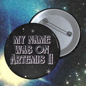 May include: A round black button with the text "MY NAME WAS ON ARTEMIS II" in white and light purple. The button has a silver pin back and is set against a starry background.