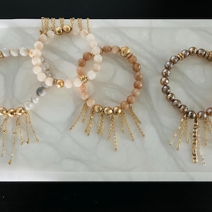 Puede incluir: Tres pulseras de cuentas con detalles dorados y abalorios de borla. Las pulseras están hechas con cuentas blancas, beige y marrones.