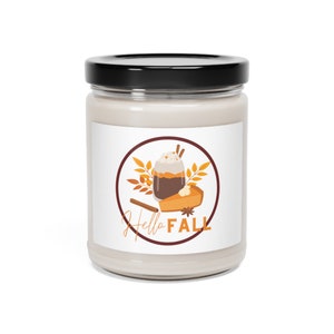Pode incluir: Uma vela de vidro em um frasco com um rótulo branco que apresenta uma ilustração de desenho animado de um latte de especiarias de abóbora e uma fatia de torta de abóbora. O rótulo também inclui o texto "Hello Fall".