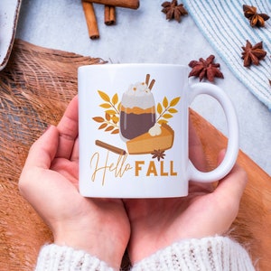 Op de afbeelding: Een witte keramische mok met een herfstthema. De mok heeft een glas koffie met slagroom en een kaneelstokje, een stuk pompoentaart en herfstbladeren. De tekst "Hello Fall" staat op de mok gedrukt.