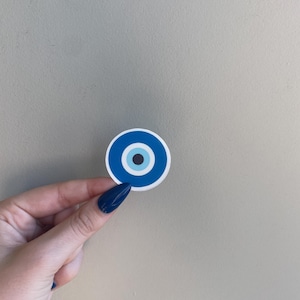 May include: A round sticker featuring a blue and white evil eye design. The sticker has a white border and a central black dot. The eye is held by a hand with blue fingernails against a neutral background.
