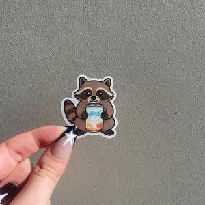 May include: A sticker featuring a cartoon raccoon holding a can of Alani Nu energy drink. The raccoon is brown and tan with a striped tail. The can is white and blue with fruit graphics.
