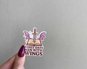 May include: A sticker with the text "I ONLY DATE MEN WITH WINGS" in a serif font. The sticker features a sword, open book, and purple wings. The sticker is held by a hand with maroon nail polish.