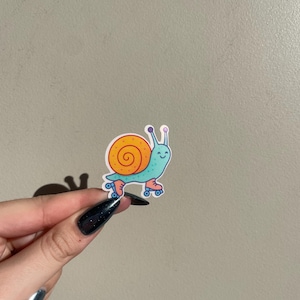 May include: A colorful sticker featuring a cartoon snail on roller skates. The snail has a blue body, an orange shell, and is wearing blue roller skates. The sticker is held up against a neutral background.