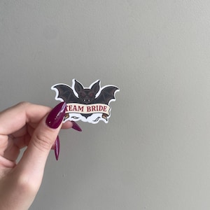 May include: A sticker featuring a black bat with red eyes and a banner that reads "TEAM BRIDE". The bat has its wings spread wide. The sticker is held up by a hand with long, dark red fingernails.