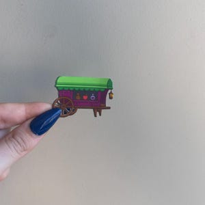 Puede incluir: Un vag&oacute;n en miniatura colorido con techo verde y cuerpo morado. El vag&oacute;n presenta una rueda de madera, peque&ntilde;os objetos decorativos y una linterna colgante. El dise&ntilde;o es caprichoso y de dibujos animados, con un enfoque en colores brillantes y detalles l&uacute;dicos.