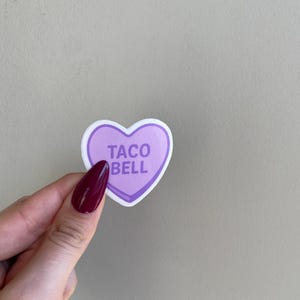 May include: A heart-shaped sticker with a purple center and white border. The text "TACO BELL" is printed in white on the purple background. The sticker is held by a hand with a burgundy nail polish.