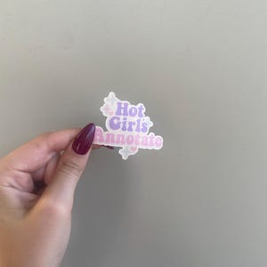 May include: A sticker with the text "Hot Girls Annotate" in a pastel color scheme. The sticker has a white border and small star accents. The sticker is held by a hand with a maroon nail polish.
