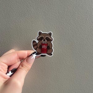 May include: A sticker featuring a cartoon raccoon holding a Dr. Pepper can. The raccoon is brown and tan with a black mask and tail. The Dr. Pepper can is red with white text.