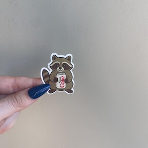 May include: A sticker featuring a cartoon raccoon holding a Diet Coke can. The raccoon is brown and white with a striped tail. The Diet Coke can is white with red text.