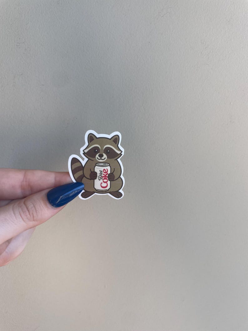 May include: A sticker featuring a cartoon raccoon holding a Diet Coke can. The raccoon is brown and white with a striped tail. The Diet Coke can is white with red text.