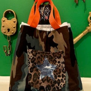 May include: A camouflage tote bag with a large pocket on the front. The pocket is made of leopard print fabric and has a blue star in the center. The bag has orange handles.