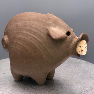 May include: A brown ceramic pig figurine with a cork in its mouth. The pig has small, rounded legs, a short tail, and small ears. The figurine is a matte finish and has a rustic appearance.