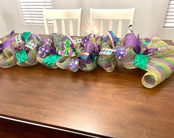 May include: A Mardi Gras themed table runner with purple, green, and gold mesh, ribbons, and decorative accents.