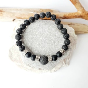 May include: Black lava rock and matte black bead bracelet with silver spacer beads. The bracelet is on a grey stone surface.
