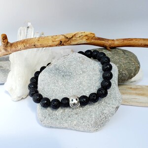 May include: Black lava rock bracelet with a silver smiley face charm. The bracelet is on a grey rock surface.