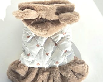 May include: A white quilted dog coat with a diamond pattern and small pink hearts. The coat has a fluffy brown faux fur collar and trim. The coat is designed to keep a small dog warm and stylish.