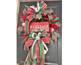 May include: A festive Christmas wreath with red and green ribbons, featuring a sign that reads "Santa's Workshop". The wreath is adorned with decorative elements and bows, perfect for holiday decor.