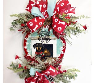 May include: A red and white Christmas wreath with a dog wearing a sweater in front of a fireplace. The wreath has a sign that says "Dear Santa I can explain".