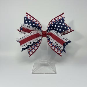 May include: A patriotic hair bow featuring a red, white, and blue color scheme. The bow has a red center with white lace trim and is adorned with a navy blue star pattern and a red and white checkered pattern. The bow is displayed on a clear stand.