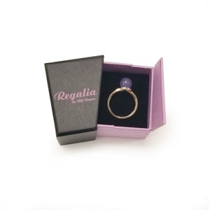 Puede incluir: Un anillo con una piedra preciosa p&uacute;rpura en una caja rosa con el texto "Regalia by Mili Ramos" en la caja.