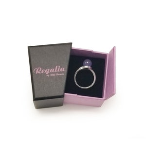 Puede incluir: Un anillo con una piedra preciosa p&uacute;rpura en una caja de joyer&iacute;a rosa y negra. La caja tiene el texto "Regalia by Ulli Ramos" impreso en el lateral.