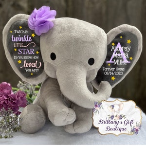 May include: A plush gray elephant toy with a purple fabric flower on its left ear. The left ear features the text Twinkle twinkle LITTLE STAR Do You Know How loved You Are? with yellow stars. The right ear displays the name Aubrey Maria Miller.