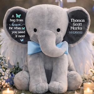 May include: A plush gray elephant with a blue bow tie and angel wings. The elephant has black eyes and gray ears. Text on the ears reads "Thomas Scott Marks 03/27/2022" and "A hug from heaven for when you need it most."