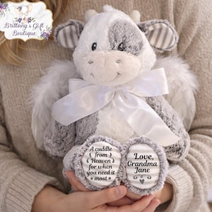 May include: A plush cow stuffed animal with white and gray fur, angel wings, and a white ribbon. The cow has text on its feet that reads "A cuddle from Heaven for when you need it most" and "Love, Grandma Jane."