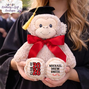 May include: A plush monkey wearing a graduation cap and a red bow tie. The monkey has the text "CLASS OF 2026" and a name printed on its feet. The monkey is light brown with a black cap and yellow tassel.