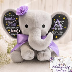 May include: A plush gray elephant toy featuring a purple fabric flower on its head and a purple satin ribbon around its neck. The ears are printed with yellow stars and text reading Twinkle twinkle little star and Aubrey Maria Miller.