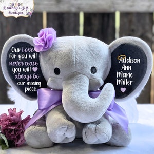 Może przedstawiać: Szary pluszowy słoń z fioletowymi skrzydłami i fioletową kokardką. Słoń ma czarne filcowe ucho z tekstem "Our Love for you will never cease you will always be our missing piece" i drugie czarne filcowe ucho z tekstem "Addison Ann Marie Miller".